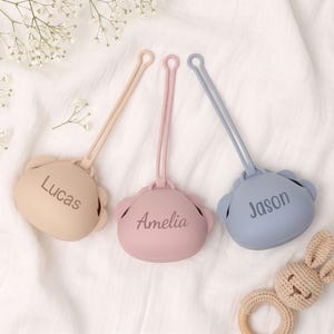 May include: Three silicone pacifier cases in beige, dusty pink, and slate blue, each featuring a long strap and a bear-shaped design. The cases are personalized with the names Lucas, Amelia, and Jason engraved in a dark font on the front surface.