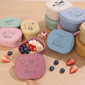 May include: A collection of square silicone snack containers in various pastel colours, each featuring a unique black line-art graphic and a personalised name. One open container holds blueberries, strawberries, and star-shaped snacks, accompanied by a small wooden spoon.