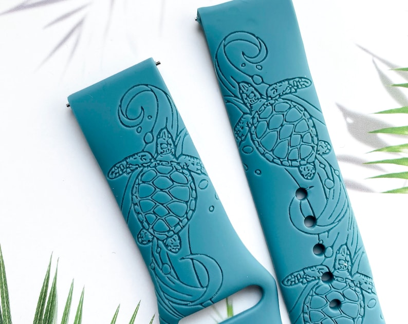 May include: Two teal blue silicone watch bands with an embossed sea turtle design. The bands have a textured surface and are designed for a smartwatch.