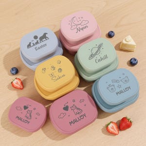 Op de afbeelding: Een verzameling rechthoekige siliconen voedselbakjes in pasteltinten zoals roze, blauw, geel en groen. Elk deksel is voorzien van een unieke grafische print en een naam, waaronder Easter, Napier, Cahill, Sabine en Malloy, naast vers fruit.