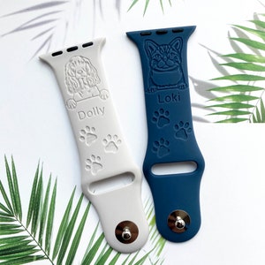 K&ouml;nnte beinhalten: Zwei personalisierte Apple Watch-Armb&auml;nder, eines wei&szlig; mit einer Hundesilhouette und dem Namen "Dolly" und das andere blau mit einer Katzensilhouette und dem Namen "Loki". Beide Armb&auml;nder haben Pfotenabdr&uuml;cke.