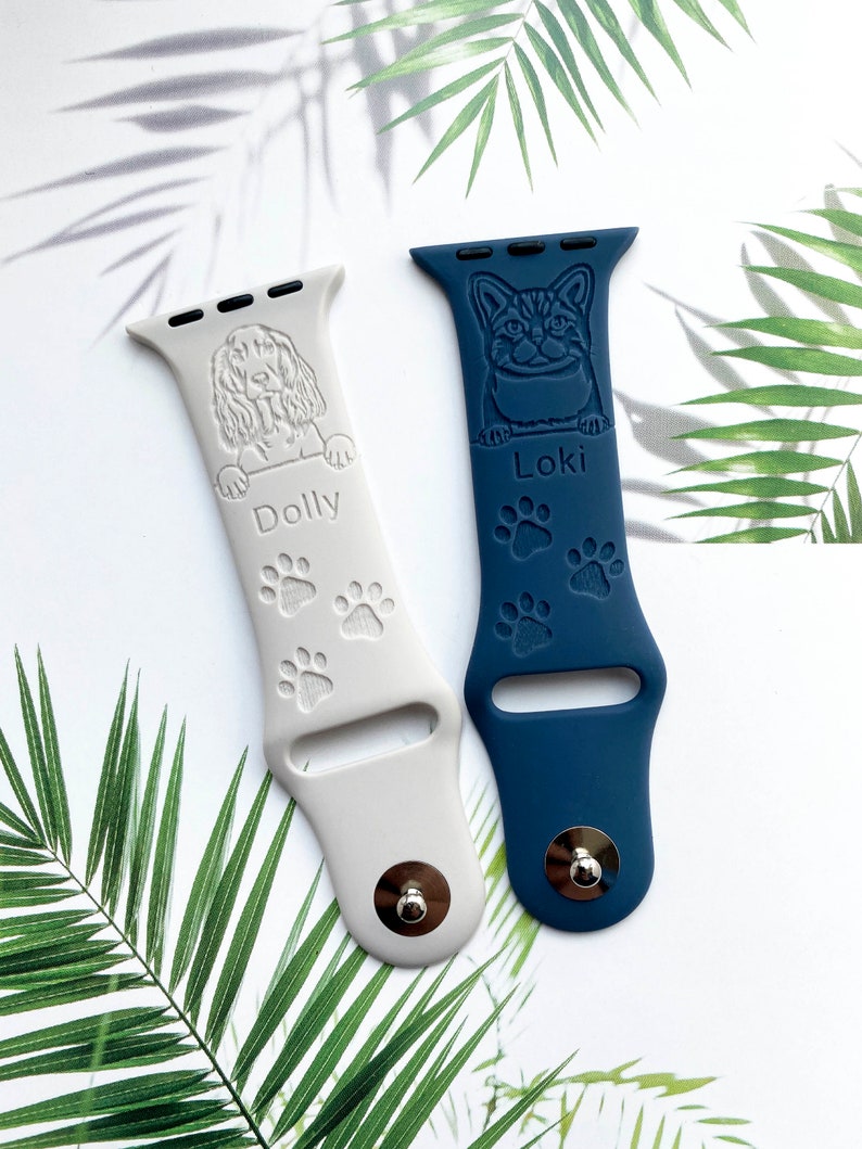 K&ouml;nnte beinhalten: Zwei personalisierte Apple Watch-Armb&auml;nder, eines wei&szlig; mit einer Hundesilhouette und dem Namen "Dolly" und das andere blau mit einer Katzensilhouette und dem Namen "Loki". Beide Armb&auml;nder haben Pfotenabdr&uuml;cke.
