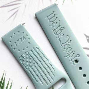 May include: Two light blue silicone watch straps with engraved designs. One strap features the text "We the People" and the other strap features a circle of stars with the year "1776" and a stylised American flag.