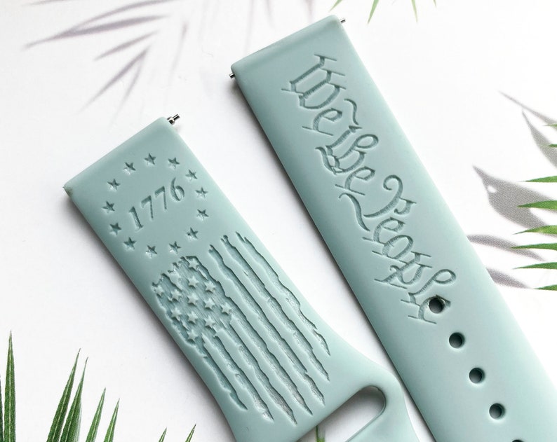 May include: Two light blue silicone watch straps with engraved designs. One strap features the text "We the People" and the other strap features a circle of stars with the year "1776" and a stylised American flag.