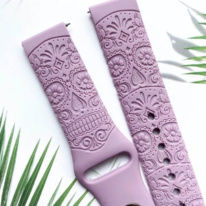 Pu&ograve; includere: Un paio di cinturini per orologio in silicone lilla con un design dettagliato di teschio di zucchero. I cinturini sono in rilievo con intricati motivi floreali e hanno una fibbia argentata.