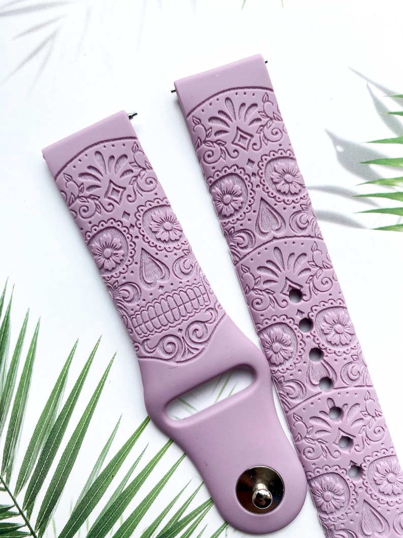 Pu&ograve; includere: Un paio di cinturini per orologio in silicone lilla con un design dettagliato di teschio di zucchero. I cinturini sono in rilievo con intricati motivi floreali e hanno una fibbia argentata.
