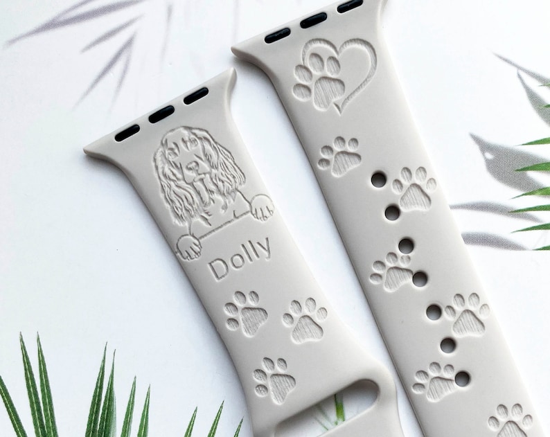 K&ouml;nnte beinhalten: Ein graues Silikonarmband f&uuml;r die Apple Watch mit einem Pfotenabdruck-Design und einem Herz mit einem Pfotenabdruck darin. Das Armband ist mit einem Hundeportrait und dem Namen "Dolly" personalisiert.