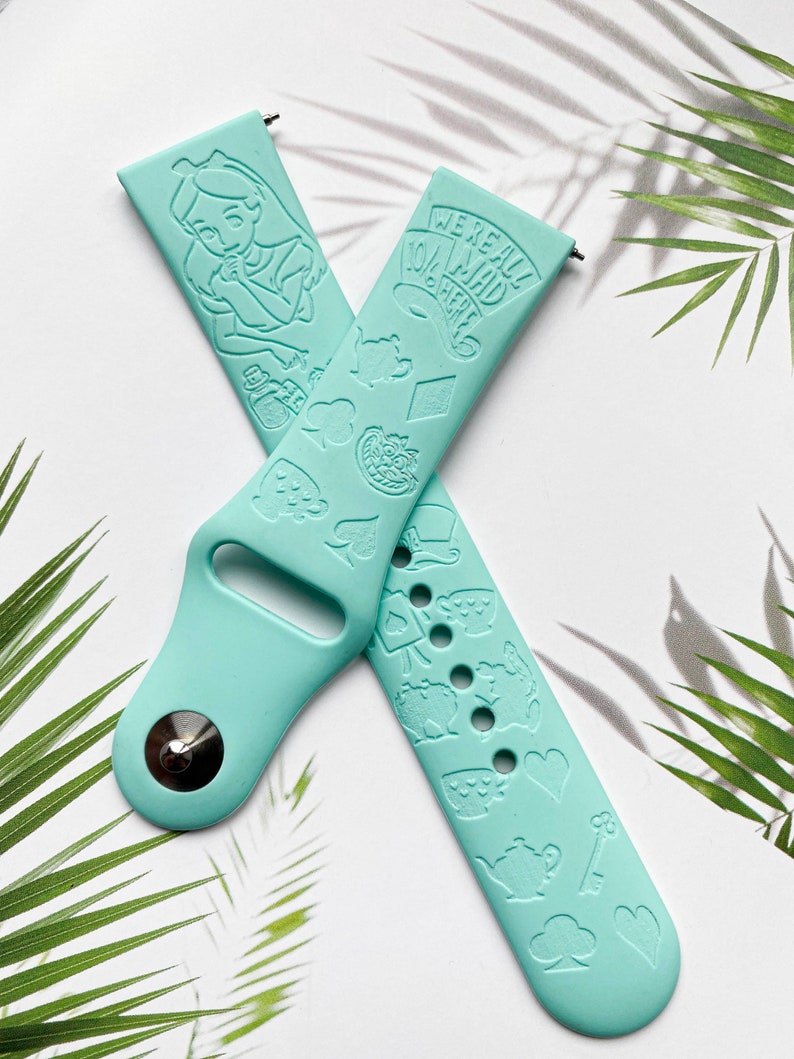 May include: A light blue silicone Apple Watch band with a white Alice in Wonderland design. The band features a variety of characters and symbols from the story, including Alice, the Mad Hatter, the Cheshire Cat, and playing cards. The text "We're all mad here" is printed on the band.