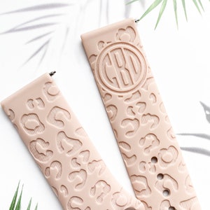 May include: Two blush pink silicone watch bands with a leopard print design. One band has a monogrammed circle with the letters "CBO" in the center.