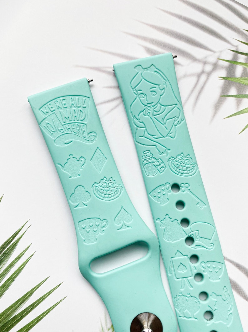 May include: Two turquoise blue silicone watch bands with a white Alice in Wonderland theme. One band features Alice, the Mad Hatter, playing cards, and a teapot. The other band features the text "We're all mad here".