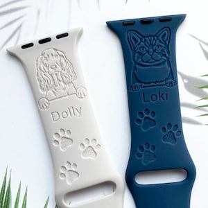 K&ouml;nnte beinhalten: Zwei personalisierte Apple Watch-Armb&auml;nder, eines wei&szlig; mit einem Hund und dem Namen "Dolly" und das andere blau mit einer Katze und dem Namen "Loki". Beide Armb&auml;nder haben Pfotenabdr&uuml;cke.