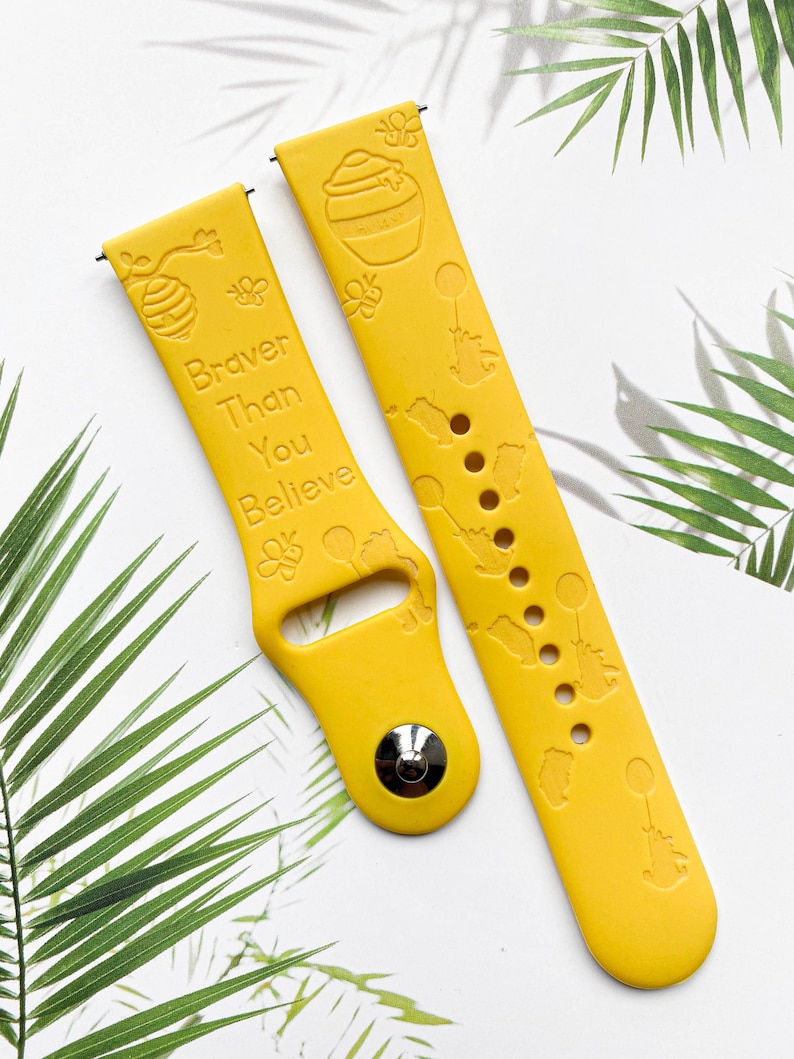 May include: Two yellow silicone watch straps with a Winnie the Pooh theme. One strap has the text "Braver Than You Believe" and a beehive design. The other strap has a honey pot and bee designs.