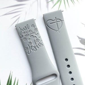 Puede incluir: Dos correas de reloj de silicona grises con diseños grabados. Una correa presenta un corazón con una cruz en su interior y la otra correa dice "Just a girl who loves Jesus" con una cruz y flores.