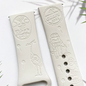May include: Two white watch bands with embossed designs. The band on the left features a house with balloons, a bird, and the text "Let's Go On An Adventure". The band on the right features a character from the movie Up, a grape soda bottle cap, and the text "Grape Soda".
