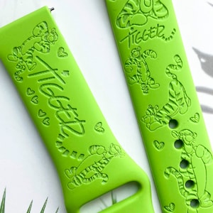 May include: Green watch band with a Tigger design. The band features a repeating pattern of Tigger's face and the word "Tigger" in a handwritten font.