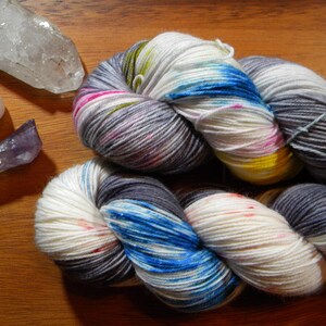 May include: A close-up of colourful yarn skeins with white, blue, grey, pink, and yellow hues. The yarn is next to several crystals, including a clear quartz, a rose quartz, and an amethyst crystal. The yarn is likely for knitting or crochet projects.