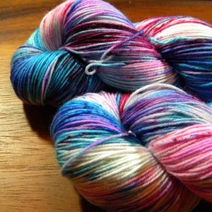 May include: Close-up of colorful yarn skeins. The yarn features a blend of blues, purples, pinks, and white. The yarn is tightly wound, showing the texture and color variations. The skeins are on a wooden surface.