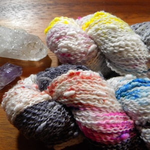 Puede incluir: Primer plano de hilo teñido a mano en colores blanco, rosa, azul, amarillo y gris. El hilo está junto a varios cristales, incluyendo cuarzo transparente y amatista. El hilo es probablemente para proyectos de tejido o ganchillo.