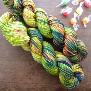 May include: Two skeins of colorful yarn with a mix of green, yellow, red, and blue hues. Several wrapped candies are scattered nearby. The yarn is likely for knitting or crochet projects.