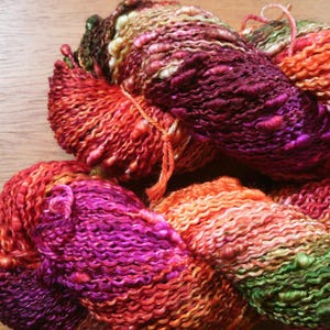 May include: A skein of hand-spun art yarn featuring a vibrant variegated color palette of deep magenta, bright orange, warm red, and olive green. The yarn has a highly textured, irregular surface with thick and thin sections and small knots.