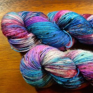 May include: Close-up of colorful yarn skeins. The yarn is a blend of blues, purples, pinks, and white, with subtle flecks of other colors. The yarn is arranged in a twisted, coiled shape, suggesting it's ready for knitting or crocheting.