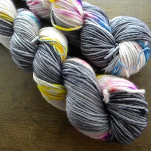 May include: Close-up of several skeins of hand-dyed yarn. The yarn is a mix of grey, white, yellow, pink, and blue. The yarn is wound into a loose, soft shape, ideal for knitting or crochet projects.