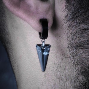 May include: A black hoop earring with a dangling, triangular, gray crystal pendant. The crystal has a multifaceted design, reflecting light. The earring is worn on the ear, showcasing a modern, edgy jewelry style.