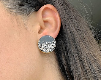 Könnte beinhalten: Ein Paar runde Ohrstecker in Schwarz und Silber mit einem goldenen Rand. Die Ohrringe sind aus Harz und haben eine glitzernde Oberfläche.