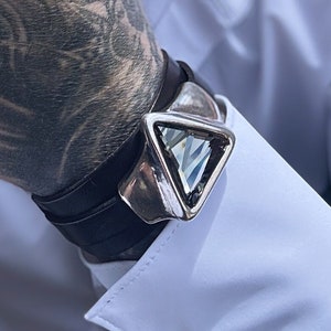 May include: A black leather bracelet with a silver triangular charm featuring a clear crystal. The bracelet is worn on a tattooed wrist.