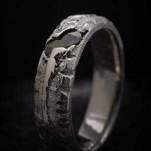 May include: Detailed shot of a silver ring with a dark gemstone. The ring features a textured, uneven surface, with a dark-coloured gemstone embedded in the band. The design suggests a handcrafted, natural aesthetic, with a focus on texture and detail.