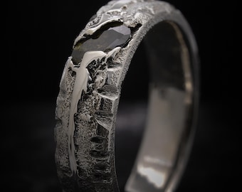 Può includere: Primo piano di un anello d'argento strutturato con una pietra preziosa scura. L'anello presenta una superficie ruvida e organica, con una pietra scura, possibilmente verde, incastonata nella fascia. Il design evoca un'estetica naturale e artigianale.