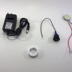 May include: A set of electrical components for a DIY project, including a black power adapter, a white LED light, a black switch, a white plastic ring, and a green and black connector.