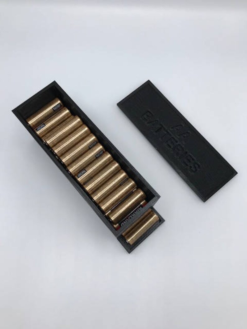 Puede incluir: Una caja de pl&aacute;stico negra con una tapa deslizante que dice "AA Batteries" en la parte superior. La caja est&aacute; abierta y llena de pilas AA de color dorado.