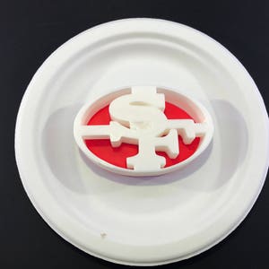 May include: A white plastic cookie cutter shaped like the San Francisco 49ers logo. The logo is on a red background.