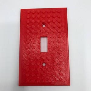 May include: A red plastic light switch cover with a textured surface that resembles Lego bricks.