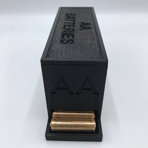 Puede incluir: Una caja de almacenamiento de bater&iacute;as de pl&aacute;stico negro con el texto "AA Batteries" en la parte superior y "AA" en la parte delantera. La caja tiene una ranura para guardar bater&iacute;as y contiene varias bater&iacute;as AA.