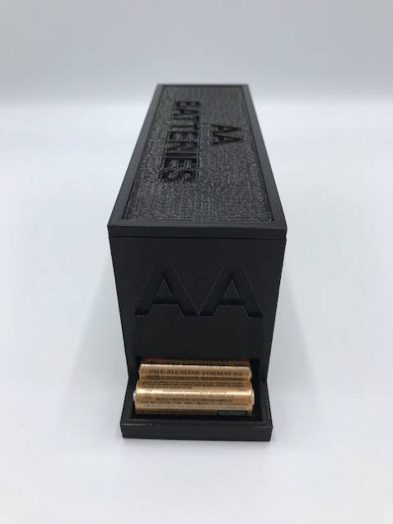 Puede incluir: Una caja de almacenamiento de bater&iacute;as de pl&aacute;stico negro con el texto "AA Batteries" en la parte superior y "AA" en la parte delantera. La caja tiene una ranura para guardar bater&iacute;as y contiene varias bater&iacute;as AA.