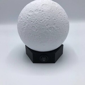 May include: A white moon-shaped lamp with a black base and an on/off switch. The lamp is designed to look like the moon with craters.