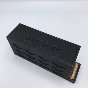 Puede incluir: Una caja negra con una superficie texturizada y el texto "AA Batteries" en la parte superior. La caja tiene una ranura en el lateral para insertar bater&iacute;as.