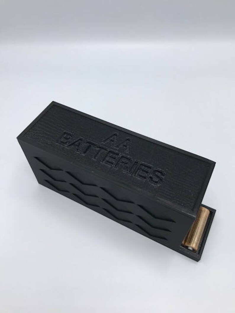 Puede incluir: Una caja negra con una superficie texturizada y el texto "AA Batteries" en la parte superior. La caja tiene una ranura en el lateral para insertar bater&iacute;as.
