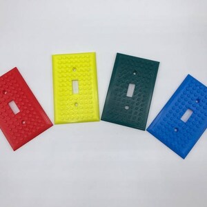 May include: Four light switch covers in different colors: red, yellow, green, and blue. Each cover has a raised texture resembling a Lego brick pattern.
