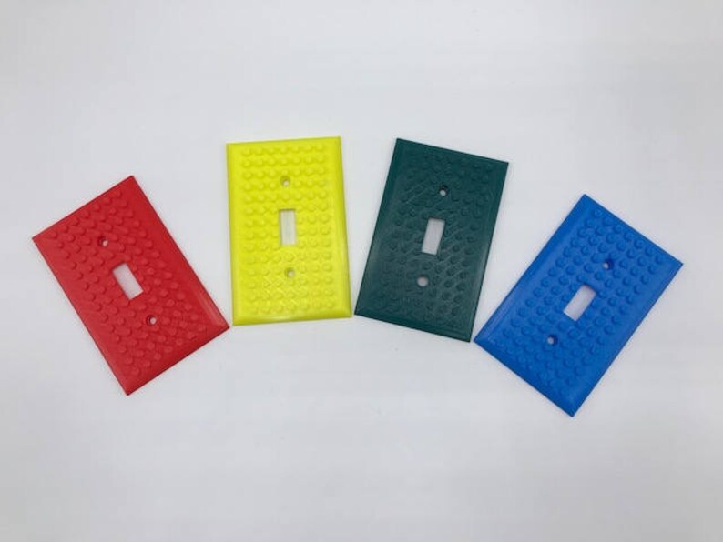May include: Four light switch covers in different colors: red, yellow, green, and blue. Each cover has a raised texture resembling a Lego brick pattern.