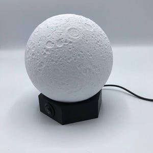 May include: A white moon-shaped night light with a black base and a power cord. The light has a textured surface that resembles the moon's surface.