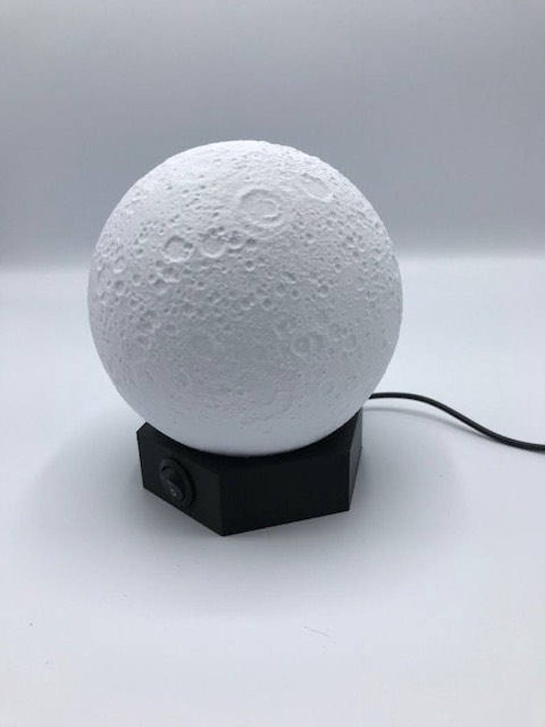 May include: A white moon-shaped night light with a black base and a power cord. The light has a textured surface that resembles the moon's surface.