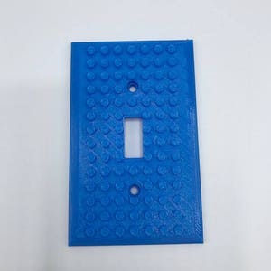 May include: A light blue plastic light switch cover with a textured surface that resembles Lego bricks.