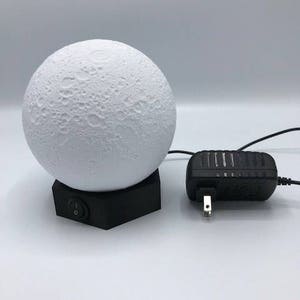 May include: A white moon-shaped lamp with a textured surface sits on a black hexagonal base. The lamp is plugged into a black power adapter with a US plug.