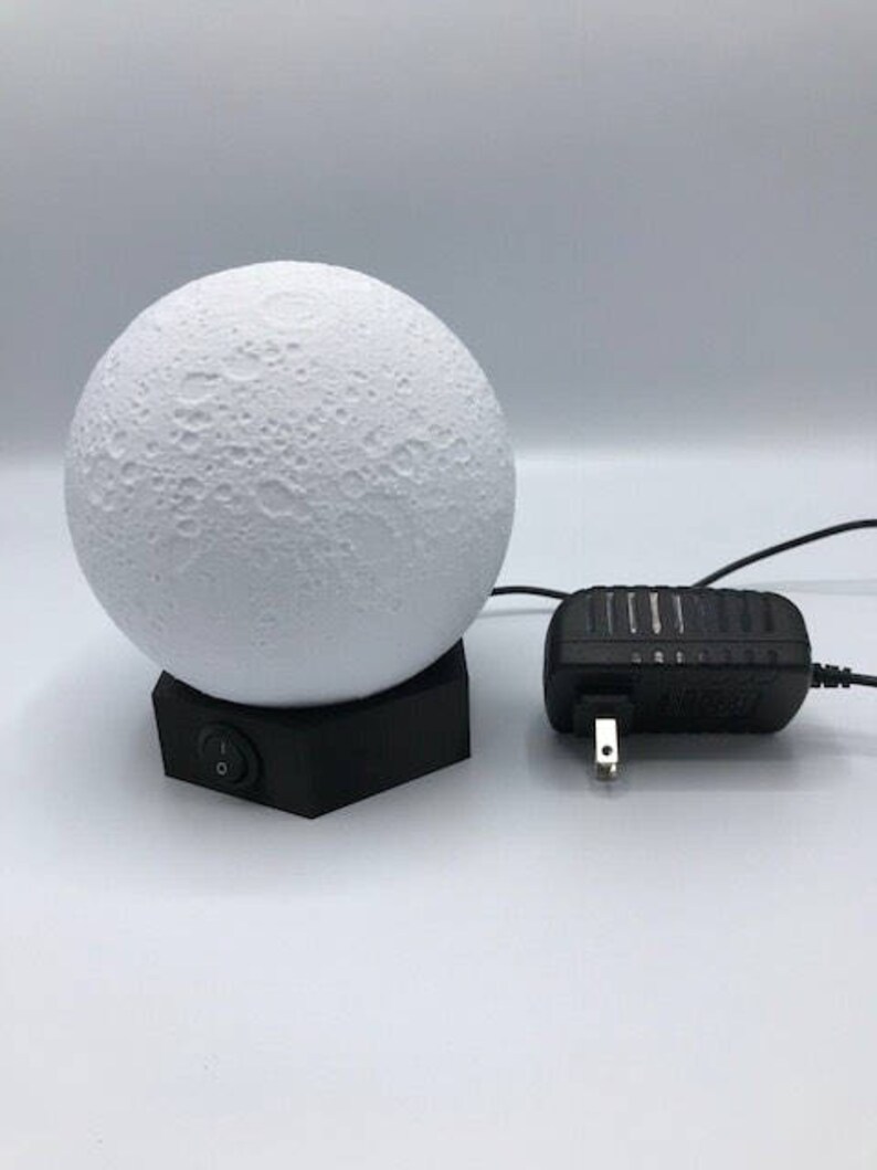 May include: A white moon-shaped lamp with a textured surface sits on a black hexagonal base. The lamp is plugged into a black power adapter with a US plug.