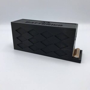 Puede incluir: Un soporte para bater&iacute;as de pl&aacute;stico negro con un patr&oacute;n de diamantes y el texto "AA Batteries" en la parte superior. El soporte tiene dos ranuras para bater&iacute;as y est&aacute; dise&ntilde;ado para mantenerlas en posici&oacute;n vertical.