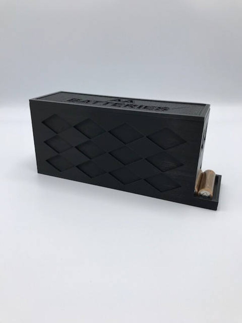 Puede incluir: Un soporte para bater&iacute;as de pl&aacute;stico negro con un patr&oacute;n de diamantes y el texto "AA Batteries" en la parte superior. El soporte tiene dos ranuras para bater&iacute;as y est&aacute; dise&ntilde;ado para mantenerlas en posici&oacute;n vertical.