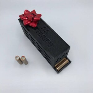 Puede incluir: Una caja negra con un lazo rojo en la parte superior. La caja dice "AA Batteries" en el lateral. Dos pilas AA est&aacute;n al lado de la caja.